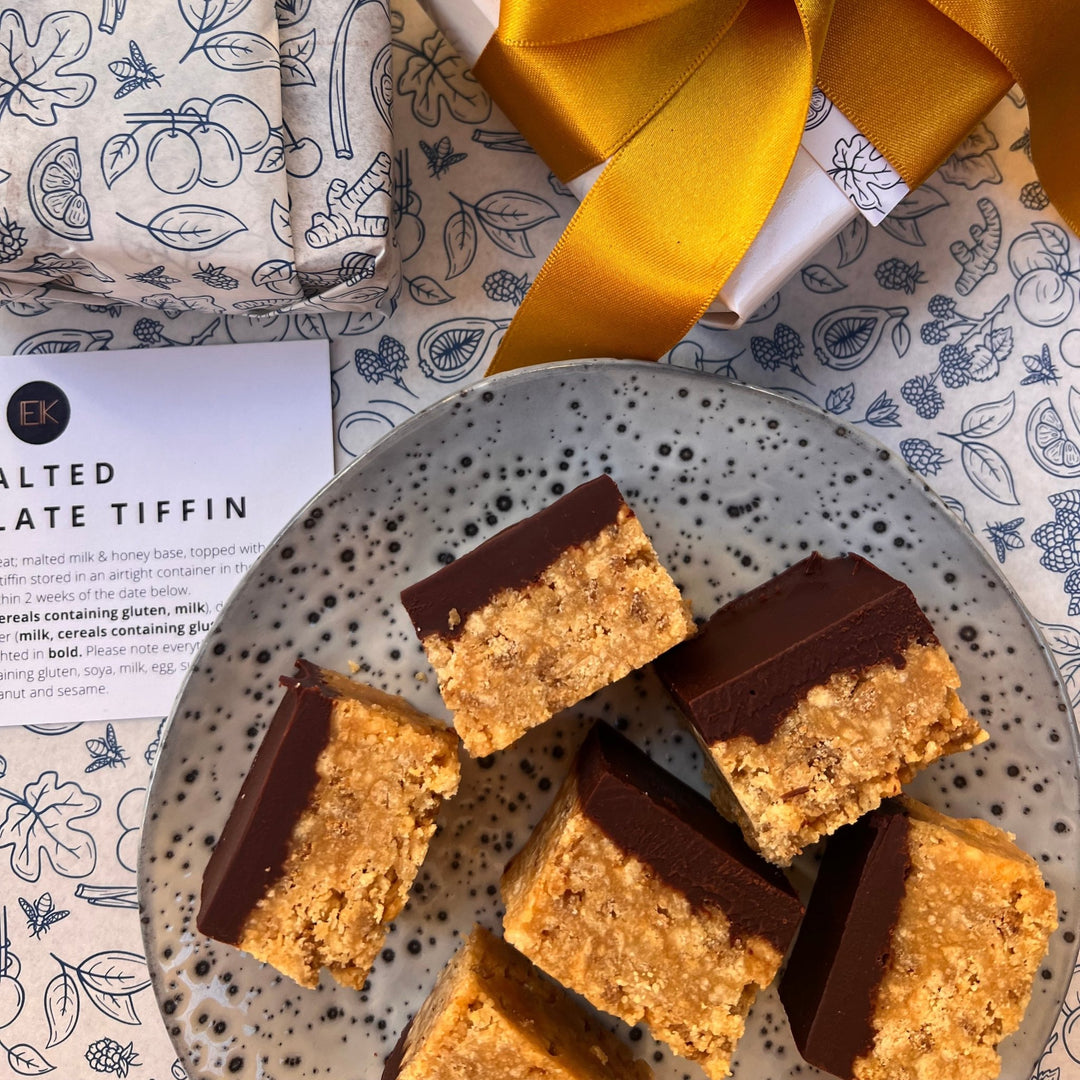 Chocolate tiffin bars arranged on a speckled plate, with a branded gift box and yellow ribbon in the background on a patterned surface.