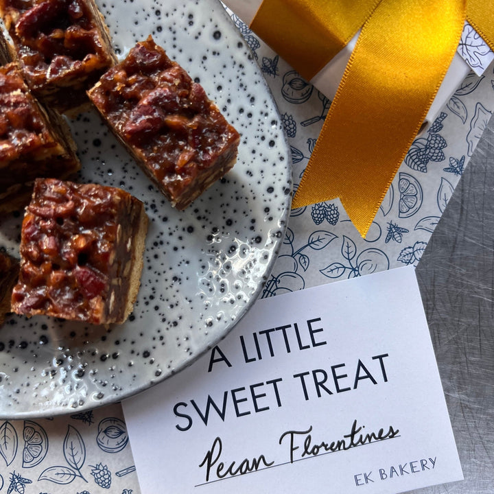 Pecan Florentines sweet treats arranged on a speckled white plate, with a white gift box wrapped in a gold ribbon and a printed card reading 'A LITTLE SWEET TREAT Pecan Florentines EK BAKERY' on a gray tabletop.