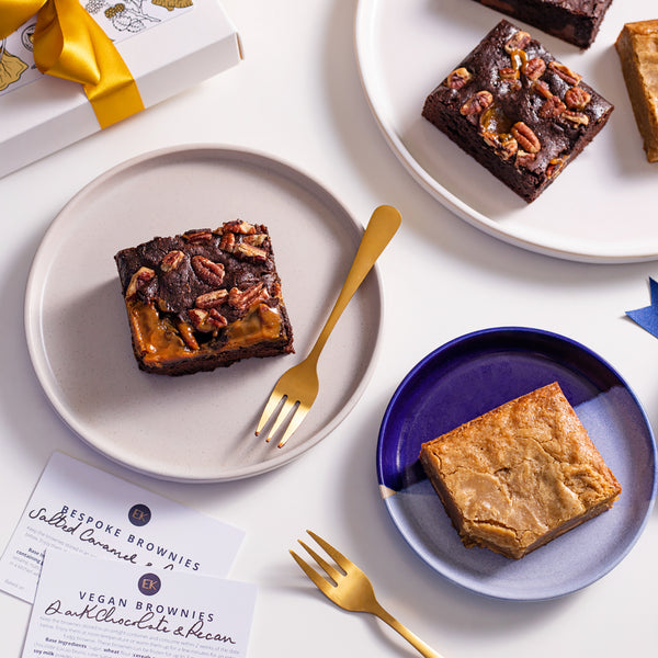 Mixed selection of brownies and blondies, set out on a table with blue and white plates and gold forks. Flavours : white chocolate blondie, salted caramel & pecan brownie